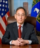 A person in a suit and tie sitting in front of flags

Description automatically generated